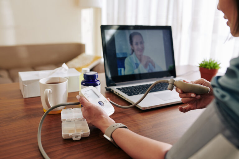 A woman uses a remotely monitored IoT blood pressure cuff while on a teleconference with her doctor.