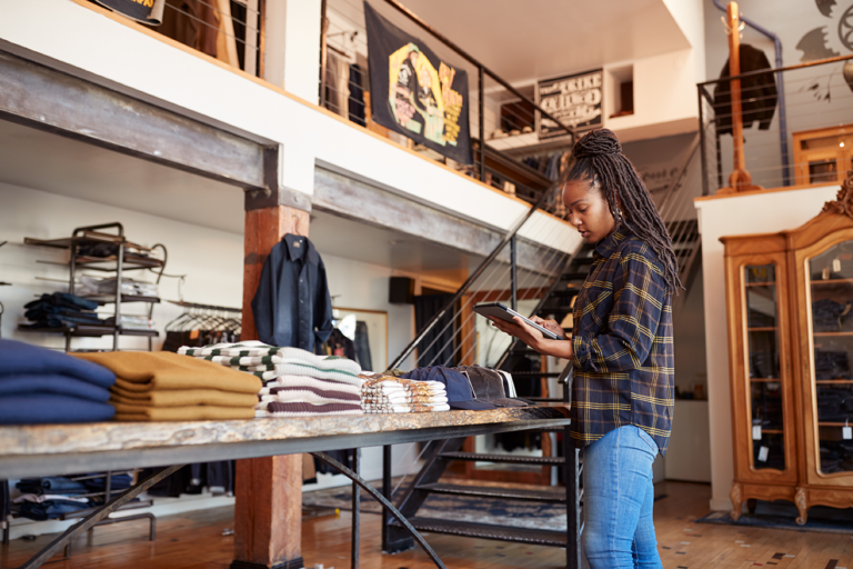 Retail Reinvented: Retail store owner using IoT-enabled tablet for inventory management in a modern boutique. Smart retail technology enhances efficiency by tracking stock levels in real-time. IoT solutions for retail improve supply chain visibility and streamline operations.